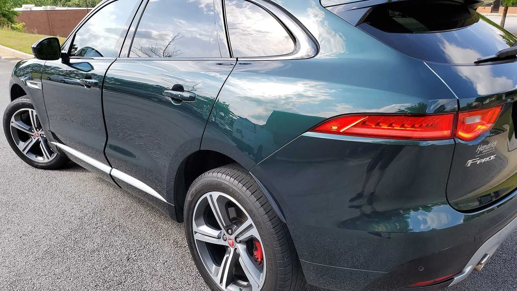 A dark green SUV with tinted windows is parked on a paved surface, showing the rear and side view with distinct red taillights and alloy wheels. The sky reflects on the vehicle's windows and body.