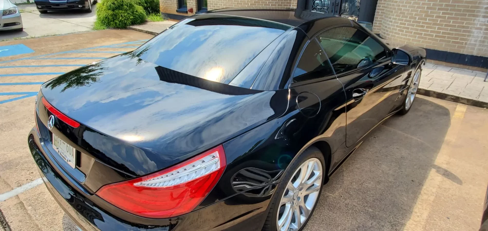 A shiny black convertible sports car is parked on a paved surface near a building. The car's roof is up, and reflections of clouds are visible on its polished surface.